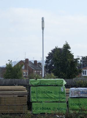 An image of a cellular communications mast, hills can be seen in the background and some terraced houses + trees can be seen too